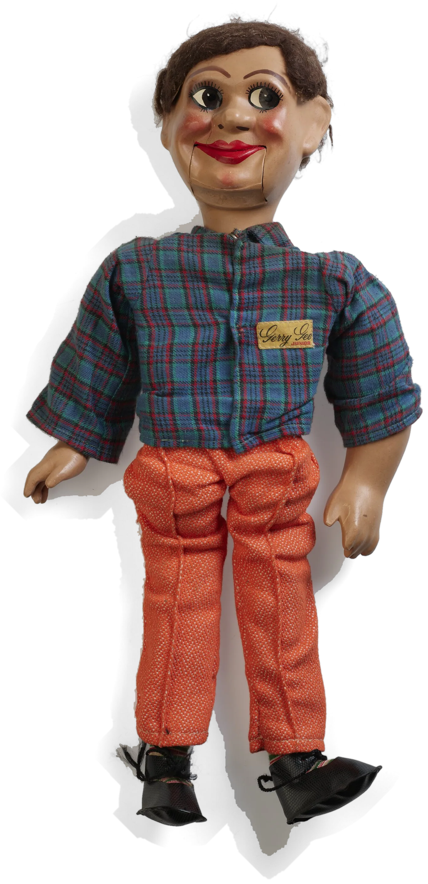 A ventriloquist’s doll standing: moulded head with brown hair and red lips, a blue and red checked shirt with a yellow Gerry Gee badge, orange trousers and black shoes.
