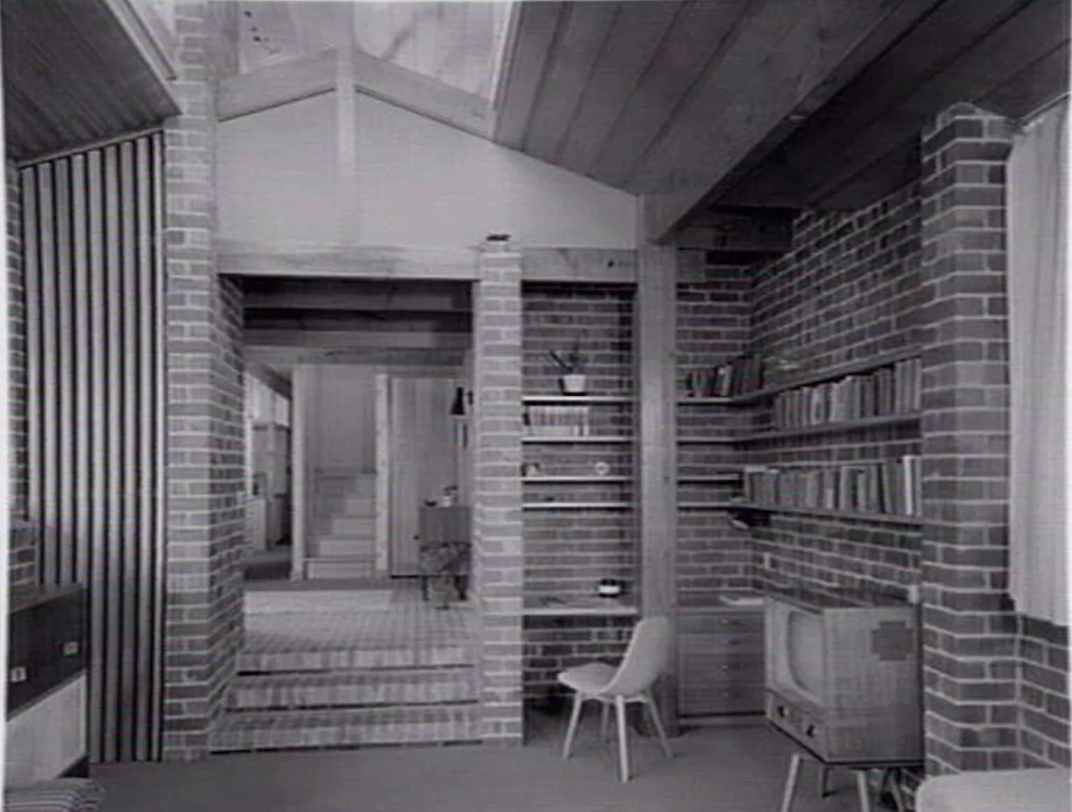 A study in a new brick house: book shelves built into the brick walls, a raked timber ceiling, a desk and a pale moulded chair, and at right a television set on legs.